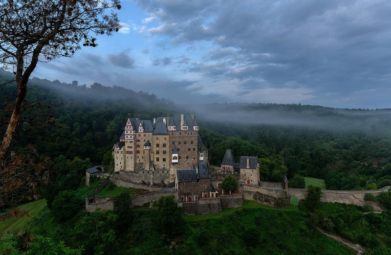 Burg Eltz mit ihren Türmen und Fachwerkerkern auf einem Felsen im bewaldeten Tal, darüber Morgennebel