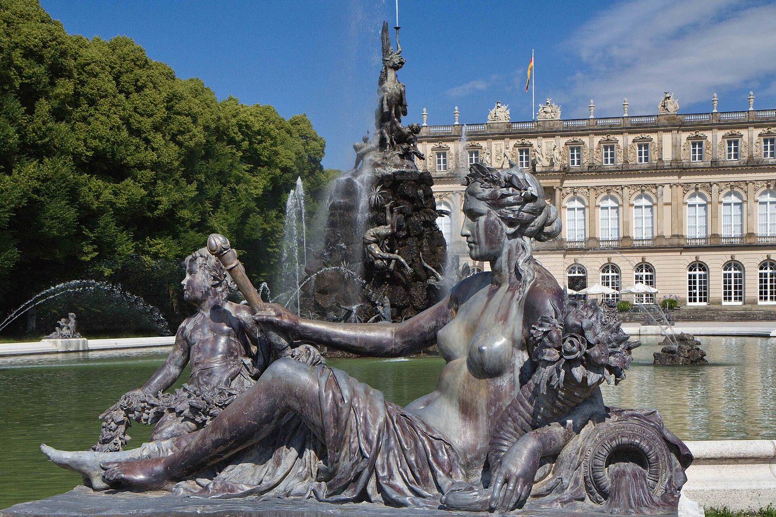 Bronzene Brunnenfigur im Schlossgarten, dahinter Fontänen und die lange Gartenfassade des Neuen Schlosses Herrenchiemsee