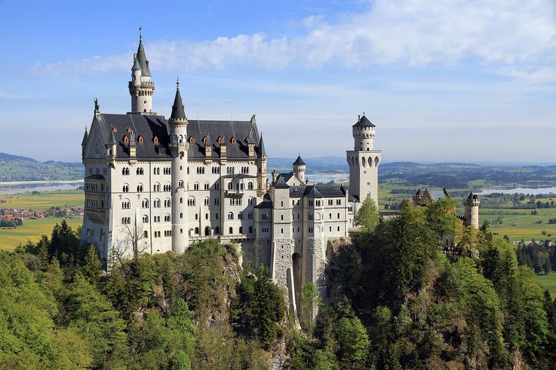 Schloss Neuschwanstein auf seinem bewaldeten Felsen, im Hintergrund das Alpenvorland mit Seen, gesehen von der Marienbrücke