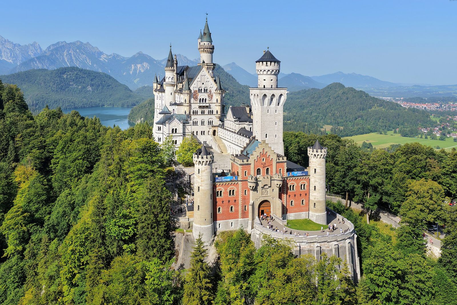 Schloss Neuschwanstein von Osten mit dem rötlichen Torbau im Vordergrund, dahinter Alpsee und Berggipfel