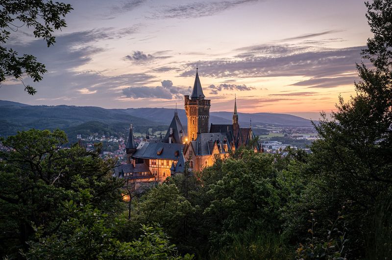 Schloss Wernigerode mit spitzen Türmen über den Baumkronen bei Sonnenuntergang, dahinter die Hügel des Harzes