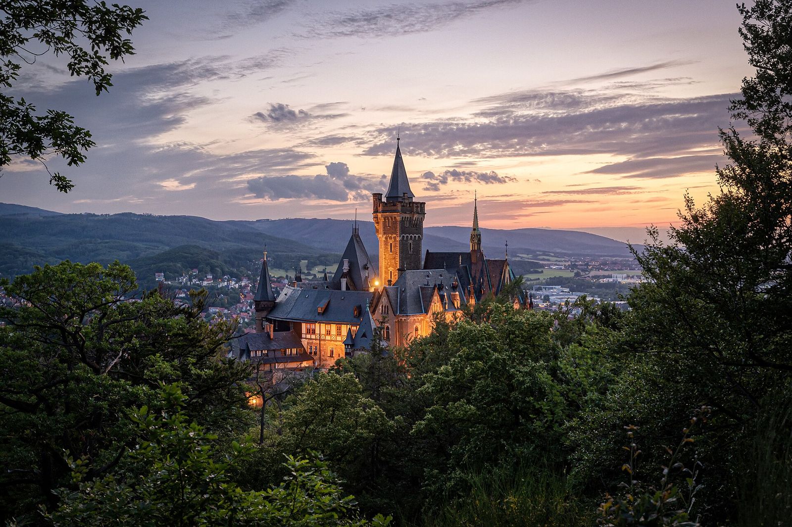Schloss Wernigerode mit spitzen Türmen über den Baumkronen bei Sonnenuntergang, dahinter die Hügel des Harzes
