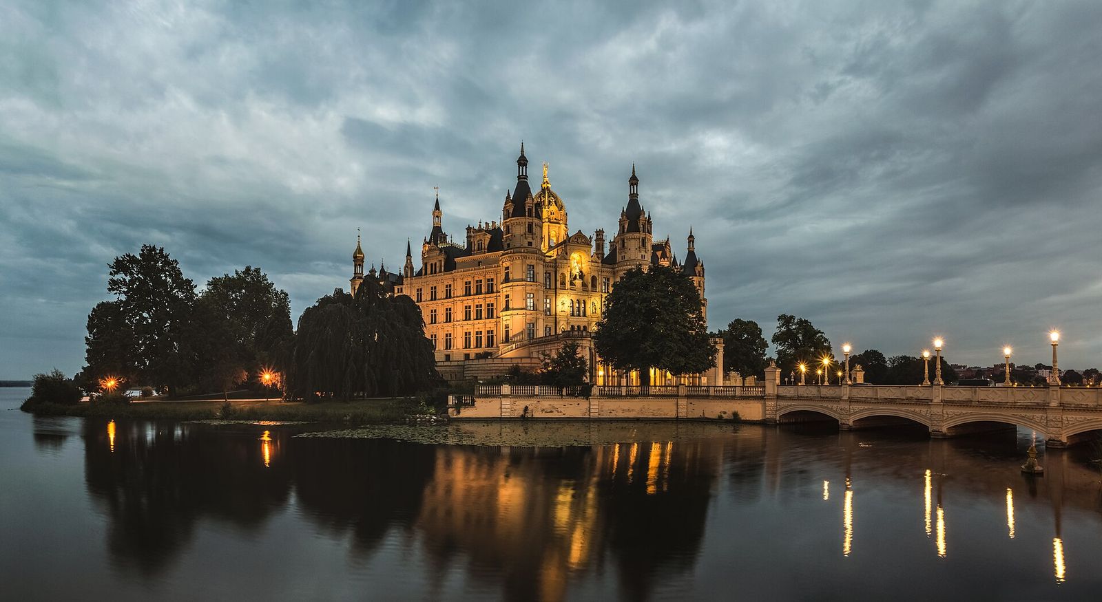 Das beleuchtete Schweriner Schloss mit goldener Kuppel in der Abenddämmerung, gespiegelt im Wasser, rechts eine Bogenbrücke